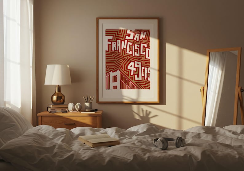 May include: A framed print featuring the words "San Francisco 49ers" in red and white. The artwork hangs above a wooden bedside table with a lamp, books, and headphones on a bed with white bedding. A mirror leans against the wall.