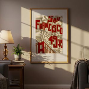 May include: A framed print with the words "San Francisco 49ers" in red block letters on a beige and white geometric design. The print is in a room with a lamp and a small potted plant.