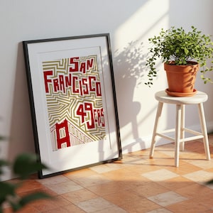 May include: Framed art print with the words "San Francisco 49ers" in red, set against a gold and white geometric pattern. The print leans against a white wall. A small potted plant sits on a white stool.
