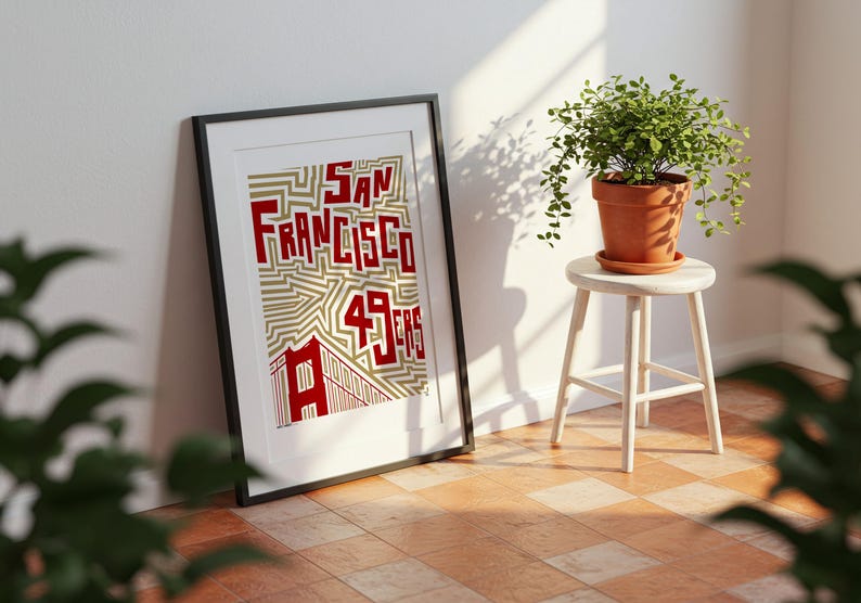 May include: Framed art print with the words "San Francisco 49ers" in red, set against a gold and white geometric pattern. The print leans against a white wall. A small potted plant sits on a white stool.