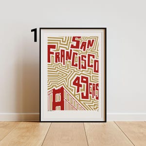 May include: Framed art print with the text "San Francisco 49ers" in red, bold lettering. The design incorporates a gold geometric pattern and a red illustration of the Golden Gate Bridge. The print is presented in a black frame.