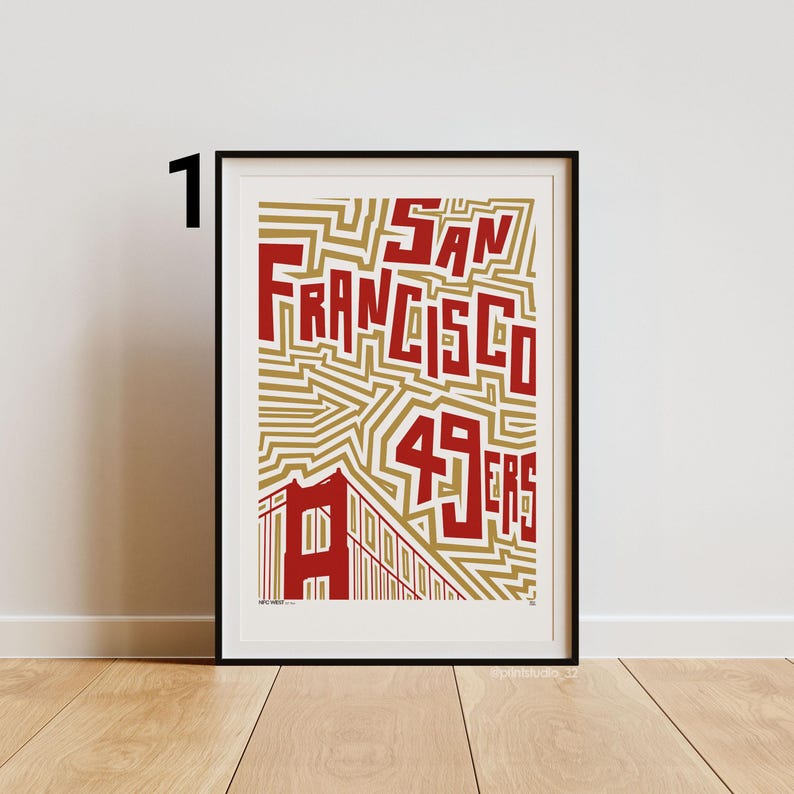 May include: Framed art print with the text "San Francisco 49ers" in red, bold lettering. The design incorporates a gold geometric pattern and a red illustration of the Golden Gate Bridge. The print is presented in a black frame.
