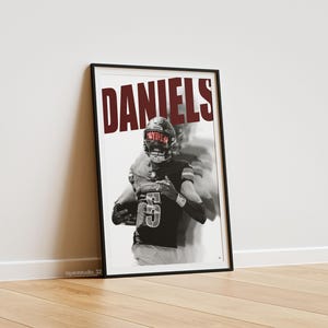 May include: Framed black and white print featuring a football player in a jersey with the number 5. The name "DANIELS" is printed in maroon at the top. The print is leaning against a white wall on a wooden floor.