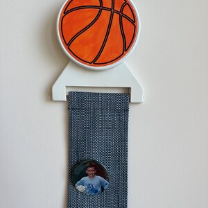 May include: A basketball-themed ribbon display. The display features a round orange basketball design with black lines, a white bracket, and a blue woven ribbon. A small circular photo is attached to the ribbon.