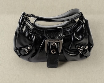 May include: A black, glossy handbag with a curved shape and a top handle. The bag features silver-toned buckles and a central strap closure. The material appears to be patent leather or a similar shiny fabric.