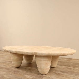 May include: A large, oval-shaped travertine coffee table with a natural beige finish. The table has four rounded, tapered legs.