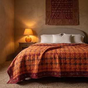 May include: A bed featuring a vibrant orange quilt with a dark red floral grid pattern, topped with white pillows. A bedside table holds a warm-toned lamp, and a dark red patterned textile hangs on the wall above the curved, light-colored headboard.