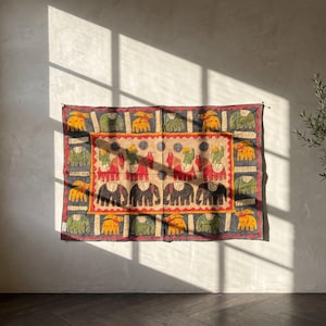 Op de afbeelding: Een rechthoekig wandkleed van textiel met een centraal paneel met rode en zwarte olifantenmotieven op een beige achtergrond, omringd door een donkergrijze rand versierd met afwisselend groene en gele olifantenfiguren in een patroon.