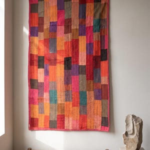 May include: A rectangular wall hanging quilt featuring a patchwork of vertical fabric strips in shades of red, orange, pink, purple, brown, and teal. The textile displays visible running stitch patterns throughout the surface.