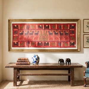 May include: A rectangular red textile wall hanging featuring embroidered animal motifs, framed in a gold-toned border. The piece displays rows of elephants and camels, centred by a stylised floral emblem, mounted above a wooden bench.