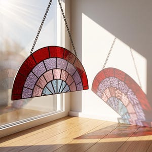 Op de afbeelding: Een halfronde glas-in-lood zonnevanger met een waaiervormig patroon van concentrische banden in rood, lichtpaars, roze en lichtblauw getextureerd glas, bijeengehouden door donkere metalen profielen en opgehangen aan een dunne zwarte metalen ketting.