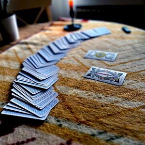 May include: A deck of playing cards fanned out on a patterned table. The cards have blue backs with a white design. Two cards are face up. A lit red candle sits in the background, creating a mystical atmosphere.