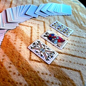 May include: A deck of playing cards with blue backs and several face-up cards. Three tarot cards are visible, featuring intricate designs and illustrations in red, blue, and gold. The cards are arranged on a textured, tan surface.