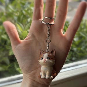 Peut inclure: Un porte-cl&eacute;s en forme de chat avec une cha&icirc;ne et un anneau argent&eacute;s. La figurine de chat est marron, blanche et beige, avec un nez rose et de grands yeux. Le chat est assis droit. Le porte-cl&eacute;s est tenu par une main.