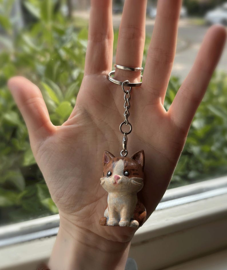 Peut inclure: Un porte-cl&eacute;s en forme de chat avec une cha&icirc;ne et un anneau argent&eacute;s. La figurine de chat est marron, blanche et beige, avec un nez rose et de grands yeux. Le chat est assis droit. Le porte-cl&eacute;s est tenu par une main.