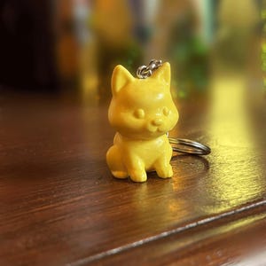 Puede incluir: Un pequeño llavero de color amarillo, impreso en 3D con forma de perro. El llavero tiene un anillo y una cadena plateados.