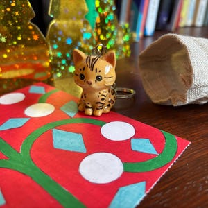 Peut inclure: Une figurine de chat marron et beige avec une bague est pos&eacute;e sur une carte rouge avec des motifs verts et blancs. Un petit sac en toile de jute ouvert et des d&eacute;corations dor&eacute;es en forme de sapin de No&euml;l sont en arri&egrave;re-plan.