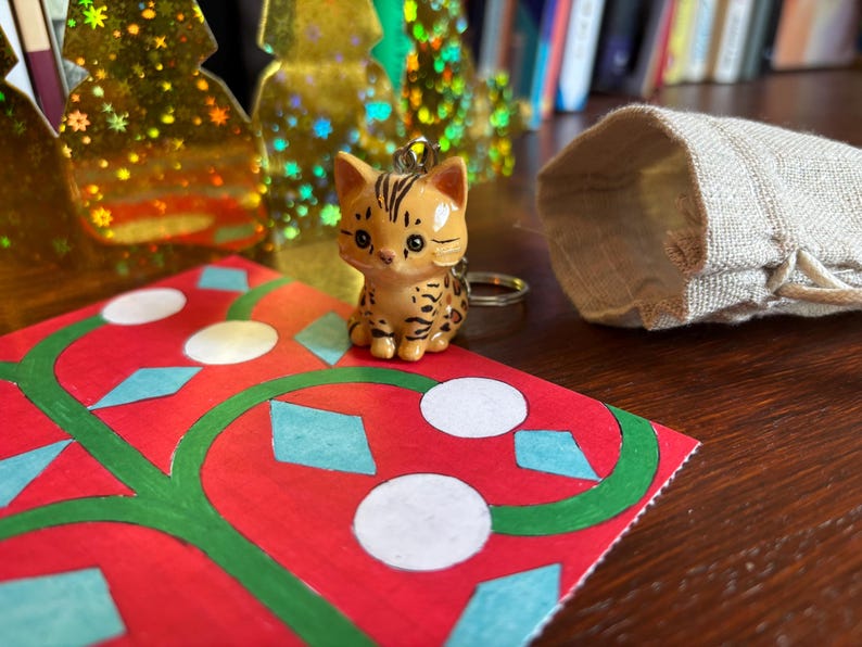 Peut inclure: Une figurine de chat marron et beige avec une bague est pos&eacute;e sur une carte rouge avec des motifs verts et blancs. Un petit sac en toile de jute ouvert et des d&eacute;corations dor&eacute;es en forme de sapin de No&euml;l sont en arri&egrave;re-plan.