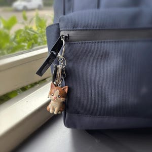 Peut inclure: Un porte-cl&eacute;s chat marron et blanc accroch&eacute; &agrave; une fermeture &eacute;clair d'un sac &agrave; dos bleu marine. Le chat est une figurine d&eacute;taill&eacute;e. Le sac &agrave; dos est en tissu textur&eacute;.
