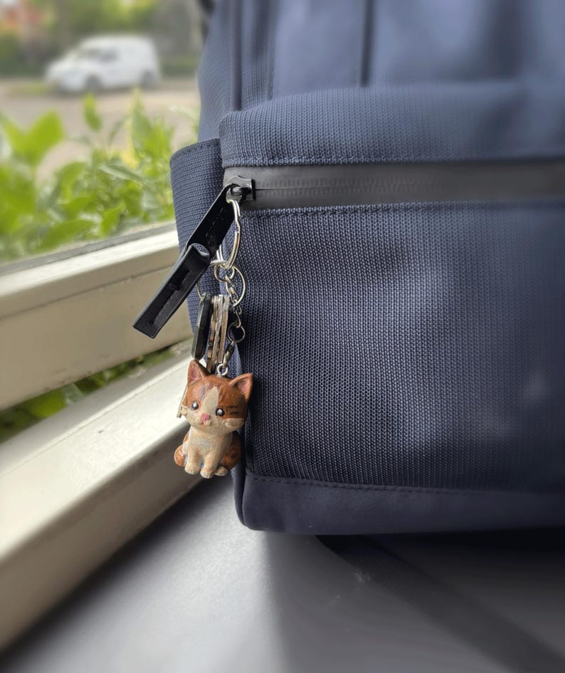 Peut inclure: Un porte-cl&eacute;s chat marron et blanc accroch&eacute; &agrave; une fermeture &eacute;clair d'un sac &agrave; dos bleu marine. Le chat est une figurine d&eacute;taill&eacute;e. Le sac &agrave; dos est en tissu textur&eacute;.