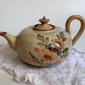 May include: A ceramic teapot with a cream-colored glaze and a hand-painted floral design featuring muted pink, blue, and green tones. The teapot includes a matching lid with a pointed finial, a curved handle, and a spout, all finished in a warm brown.