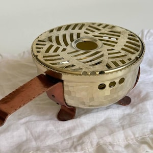 May include: A round metal fondue burner featuring a hammered gold-tone finish, a perforated top grate with a circular centre opening, and a copper-coloured handle and three small feet. The burner has a series of small circular vents along the side.