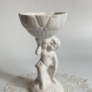 Może przedstawiać: Biała ceramiczna misa na postumencie z wyrzeźbioną figurką cherubina podtrzymującą czaszę. Misa posiada falisty brzeg z wytłoczonymi wzorami liści, a podstawę stanowi figurka klęcząca na skalistym wzniesieniu, wykończona błyszczącym szkliwem.