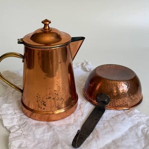Könnte beinhalten: Eine polierte Kaffeekanne aus Kupfer mit Messinggriff und passendem Deckel, neben einer kleinen, runden, gehämmerten Kupferschale mit einem dunklen, flachen Metallgriff, der seitlich vom abgerundeten Gefäß absteht.