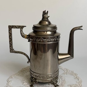 Puede incluir: Una cafetera de metal plateado con cuerpo cilíndrico, un pico largo y curvado, y un asa con patrón geométrico. La tapa está rematada con un pequeño adorno en forma de ave, y la base se apoya sobre tres patas ornamentadas con bandas decorativas.