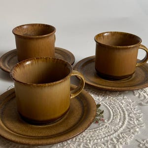 May include: Three brown ceramic coffee cups and saucers. The cups have a rounded shape with a handle, and the saucers are circular. The set has a warm, earthy tone, with variations in the glaze. The cups and saucers are arranged on a white lace tablecloth, creating a vintage feel.