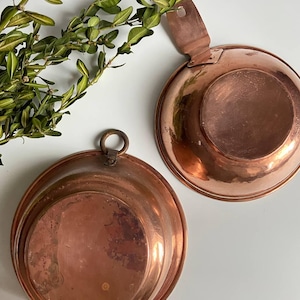 Könnte beinhalten: Zwei Kupferschalen mit glänzend rotbraunem Finish. Eine Schüssel hat einen Griff, die andere einen Ring zum Aufhängen. Grüne Blattzweige sind im Hintergrund. Diese Schalen sind wahrscheinlich für den Küchengebrauch.