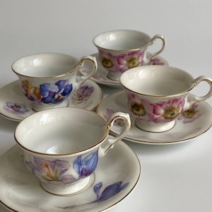 Puede incluir: Cuatro tazas de té y platillos blancos con diseños florales. Las tazas tienen ribetes y asas doradas. Los diseños florales incluyen flores moradas, rosas y amarillas. Los platillos también presentan diseños florales a juego.