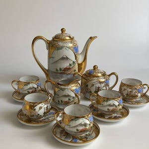 Peut inclure: Service à thé en porcelaine vintage comprenant une théière, une crémière, un sucrier et six tasses avec soucoupes. L'ensemble présente une bordure dorée et des scènes de paysage peintes à la main avec une montagne, des arbres et des accents bleus.