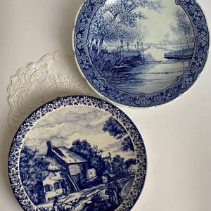 May include: Two blue and white decorative plates. One plate features a landscape scene with a river and trees. The other plate depicts a rural scene with a house and figures. Both plates have a detailed border design.