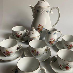 Puede incluir: Un juego de té de porcelana blanca con diseños florales y de mariposas. El juego incluye una cafetera, azucarero, lechera y varias tazas de té con platillos. Los bordes de cada pieza están adornados con detalles dorados, creando una estética elegante.