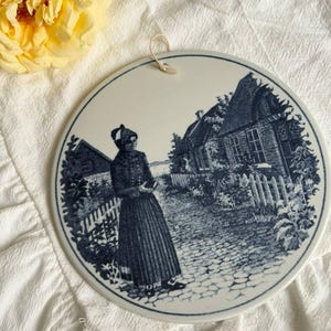 Può includere: Una placca da parete rotonda in ceramica con un'illustrazione in stile blu e bianco che raffigura una persona in abiti tradizionali in piedi su un sentiero acciottolato vicino a una casa con staccionata, dotata di un piccolo gancio per appenderla.