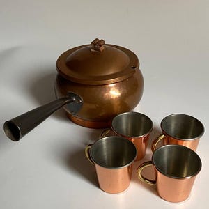 Puede incluir: Una cacerola de cobre con tapa y un mango largo y oscuro, acompañada de cuatro pequeñas tazas de cobre con asas doradas. El juego está hecho de cobre y acero inoxidable, y probablemente sea para uso en la cocina.
