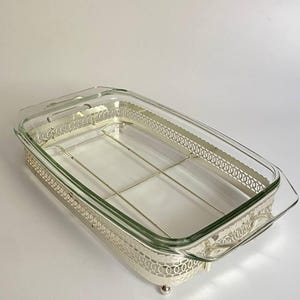 Può includere: Una pirofila rettangolare in vetro trasparente inserita in una cornice di metallo color argento. La cornice metallica presenta un motivo decorativo a filigrana sui lati, include due manici integrati e poggia su quattro piccoli piedini sferici.