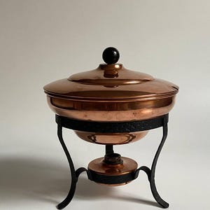 Könnte beinhalten: Ein kupferfarbenes Chafing Dish mit Deckel und einem schwarzen Schmiedeeisenständer. Das Gericht hat eine runde Form mit einem schwarzen Knopf oben. Der Ständer hat gebogene Beine und einen kreisförmigen Sockel.