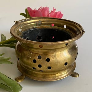 Puede incluir: Un quemador de incienso de latón vintage con un cuerpo redondo y pequeños pies. El quemador tiene numerosos agujeros pequeños para la ventilación y un asa. Una flor rosa y hojas verdes están en el fondo.