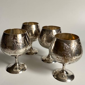 Può includere: Quattro bicchieri da brandy color argento con decorazioni floreali e a volute ornate. Ogni bicchiere ha una coppa arrotondata, un gambo corto e una base circolare. I bicchieri sono disposti su una superficie bianca.
