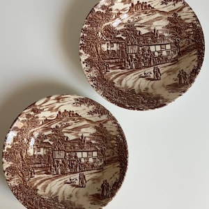 Könnte beinhalten: Zwei runde Keramikteller mit einem detaillierten braun-weißen Transferdruck-Muster. Das Design zeigt eine ländliche Szene mit einer Pferdekutsche, Personen auf einem Weg, einem Fachwerkhaus und einer Burg auf einem Hügel im Hintergrund.