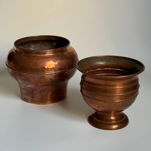 May include: Two copper vessels with a metallic finish. The larger vessel has a wide, rounded body and a straight rim. The smaller vessel features a pedestal base, a rounded bowl, and three horizontal ridges encircling the upper portion of the body.