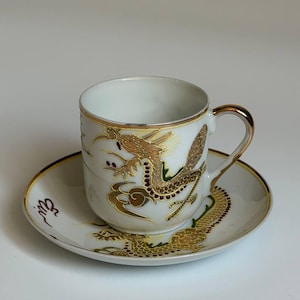 Op de afbeelding: Een witte porseleinen kop en schotel set met een goudkleurig drakenmotief en rode accenten. De kop heeft een goudkleurige rand en oor, terwijl de schotel een bijpassend drakenontwerp toont dat rond de basis van de kop loopt.