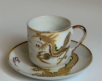 Può includere: Un set composto da tazza e piattino in porcellana bianca con un motivo a drago color oro e dettagli rossi. La tazza presenta un bordo e un manico dorati, mentre il piattino mostra un disegno di drago abbinato che circonda la base.