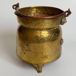 Op de afbeelding: Een koperen pot met een ronde vorm, drie decoratieve pootjes en een koperkleurige metalen zwenkhandgreep. Het buitenoppervlak is voorzien van gedetailleerde gegraveerde bloemmotieven, cirkelvormige patronen en een band met tekst.