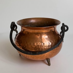 Puede incluir: Una olla de cobre martillado con cuerpo redondeado, una abertura ancha y tres pies pequeños. La olla cuenta con un asa de metal oscuro y curvada unida a dos anillos laterales. La superficie exterior tiene un acabado texturizado con hoyuelos.