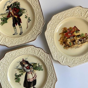 Könnte beinhalten: Drei dekorative Vintage-Teller mit gewellten Rändern und goldenem Rand. Jeder Teller zeigt eine andere Szene: einen Mann, der ein Bündel trägt, ein Gebäude mit einem Schild und eine Frau in traditioneller Kleidung. Die Teller haben einen cremefarbenen Hintergrund.