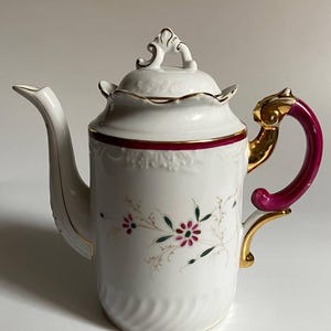 May include: White porcelain teapot with a floral design, a burgundy band, and gold accents. The handle is a deep pink color. The lid has a decorative handle and gold trim. The spout is long and slender.