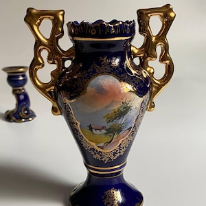 May include: A navy blue ceramic vase with gold accents and handles. The vase features a painted landscape scene and ornate gold detailing. A smaller matching candle holder is visible in the background.